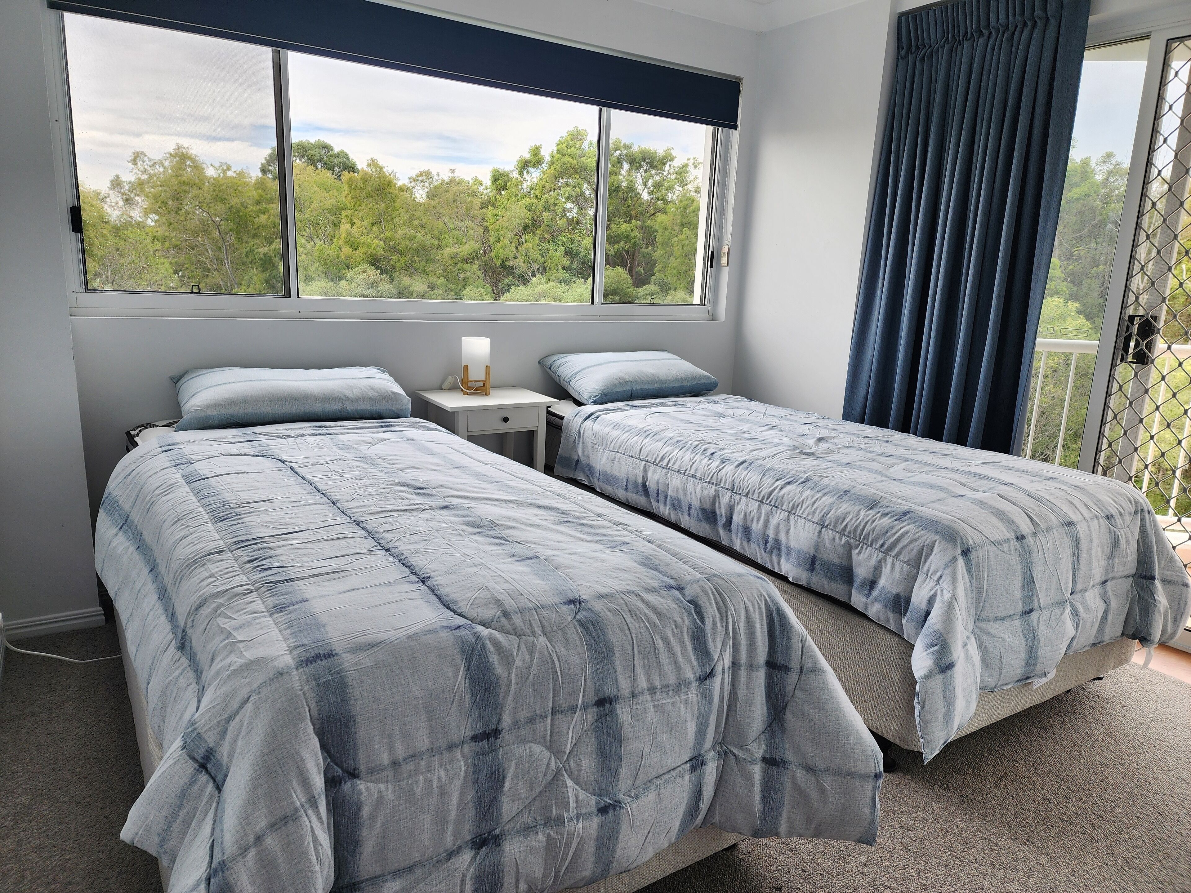 A bedroom with two beds, a nightstand with a lamp, and a view of trees through a window.
