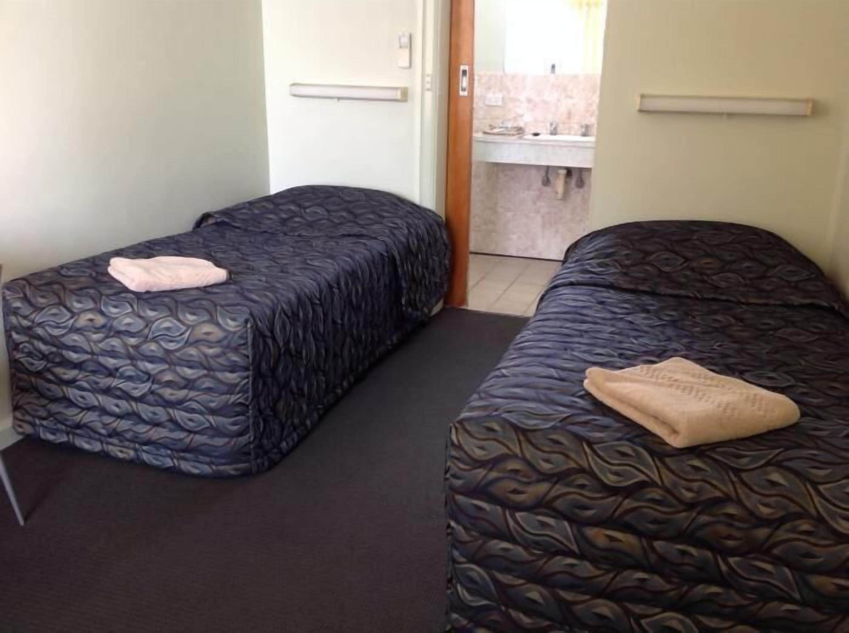 A room with two beds, each with a patterned bedspread and a folded towel. There is a door leading to a bathroom with a sink and mirror.