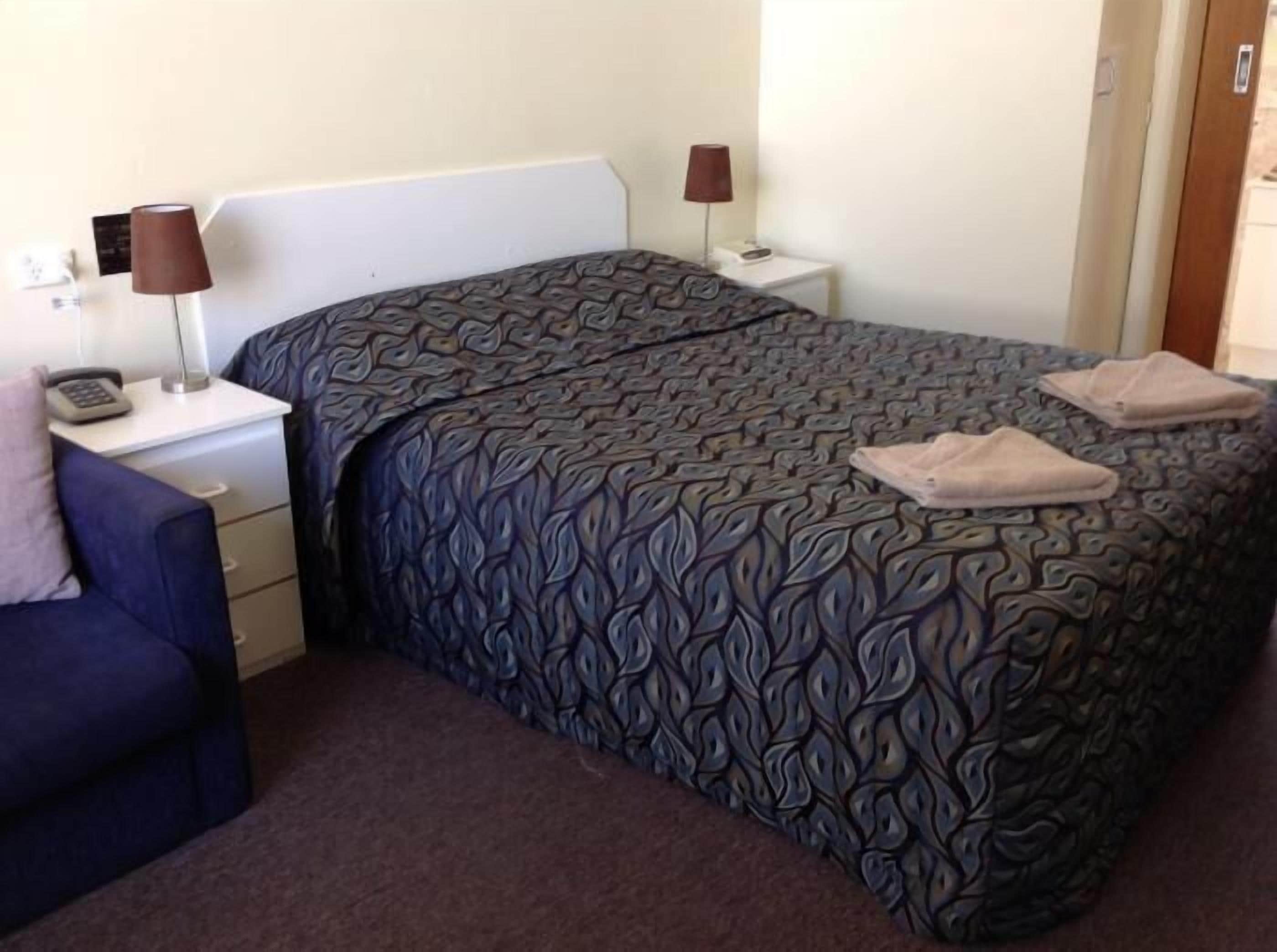 A hotel room with a patterned bedspread, a blue chair, two bedside tables with lamps, and a telephone on one of the tables.