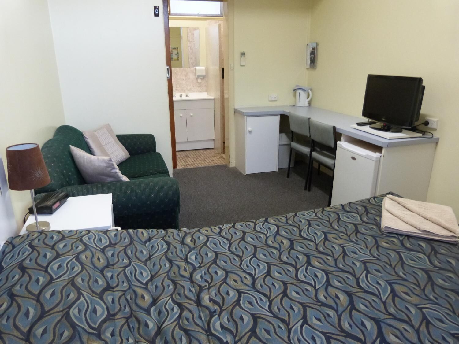 A hotel room with a bed, a desk with a computer, a chair, a small kitchenette, and a sofa.