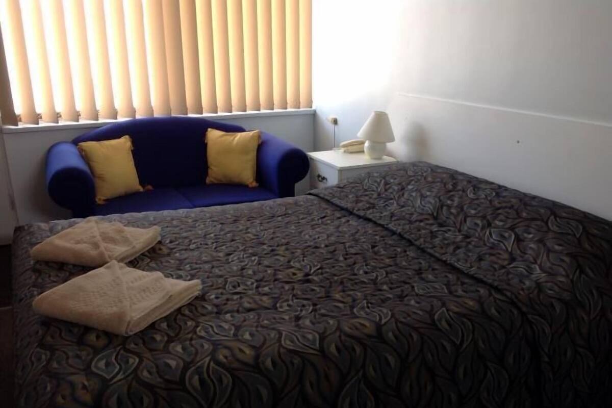 A room with a bed, a blue sofa with yellow cushions, a bedside table with a lamp, and a window with blinds.