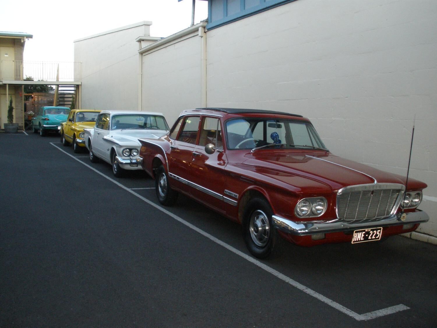 A row of vintage cars parked in a lot with a white building in the background.