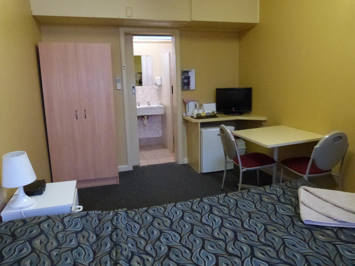 Quarto de hotel compacto com uma cama, uma escrivaninha com cadeira, televisão e uma kitchenette pequena.