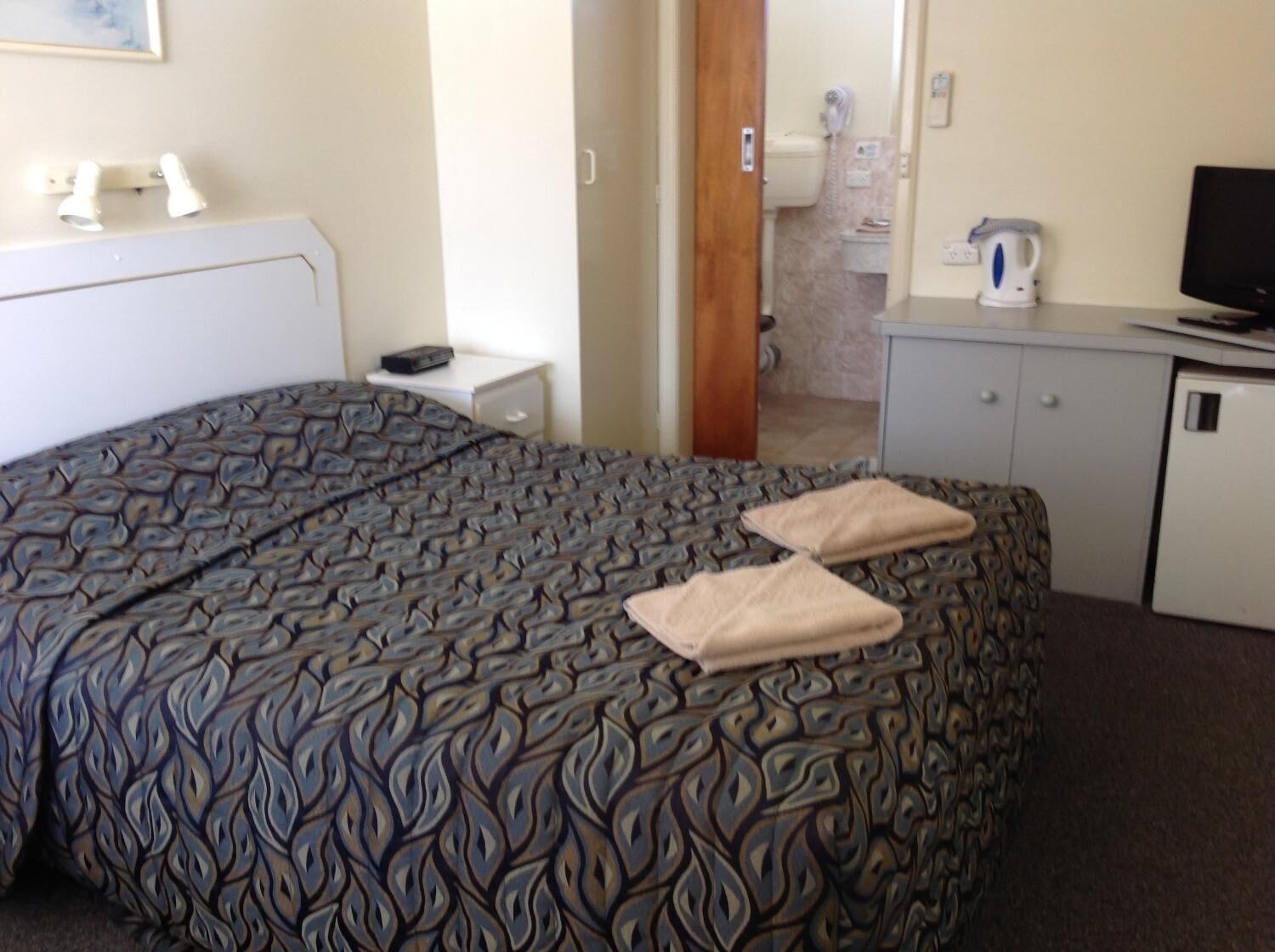 A hotel room with a patterned bedspread, a bedside table with a remote control, a television on a cabinet, and a bathroom visible through an open door.