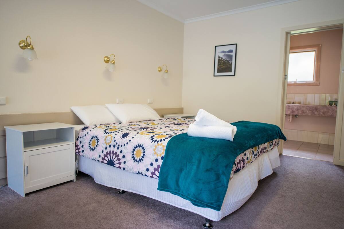 Un dormitorio con cama, mesita de noche, lámpara de pared e imagen colgada en la pared.