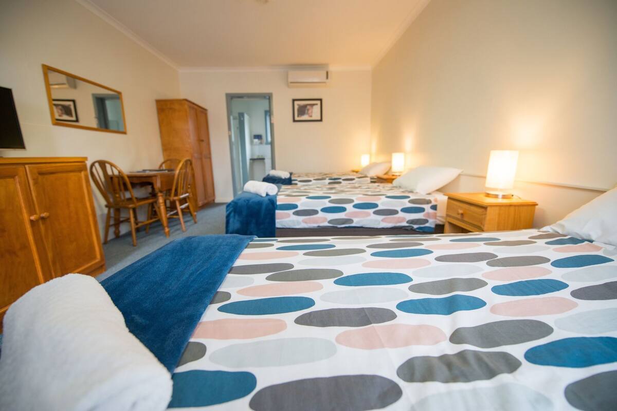 Habitación de hotel con tres camas, un escritorio, una silla, un armario y un espejo.