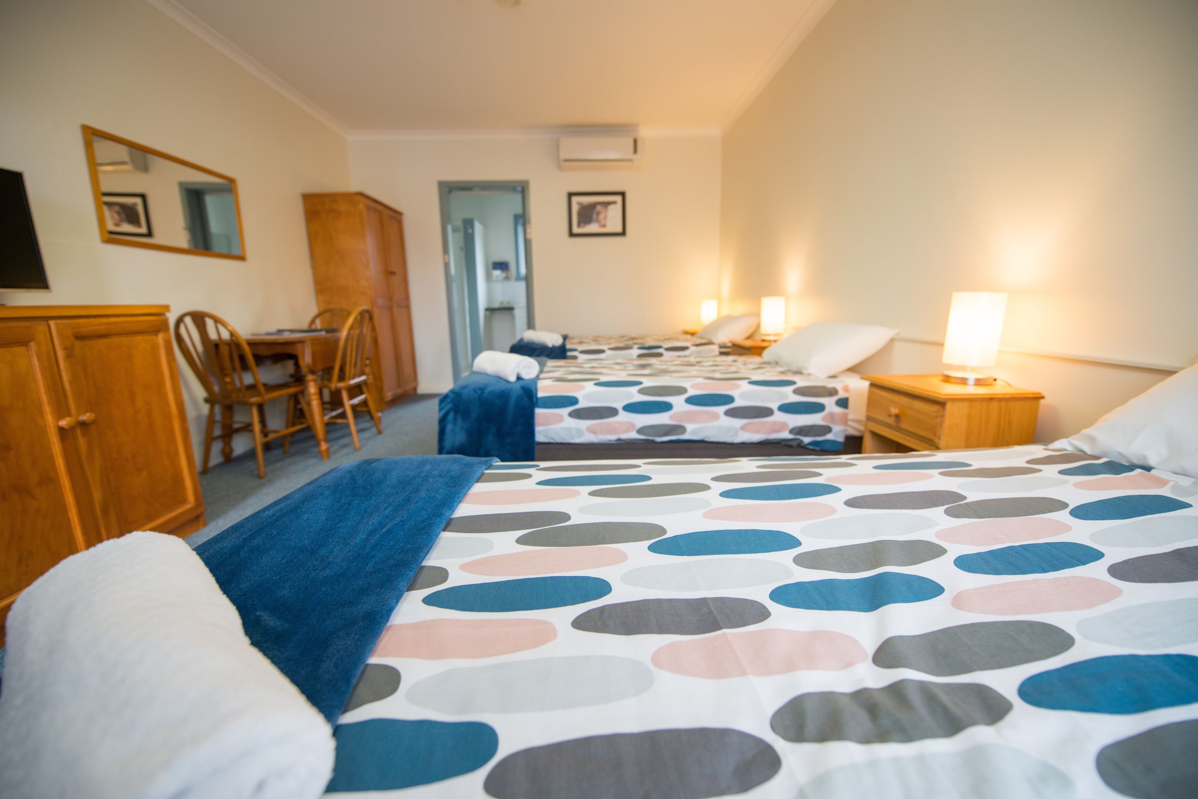 A hotel room with a long bed, a desk, and a chair. There is a wardrobe and a mirror on the wall.