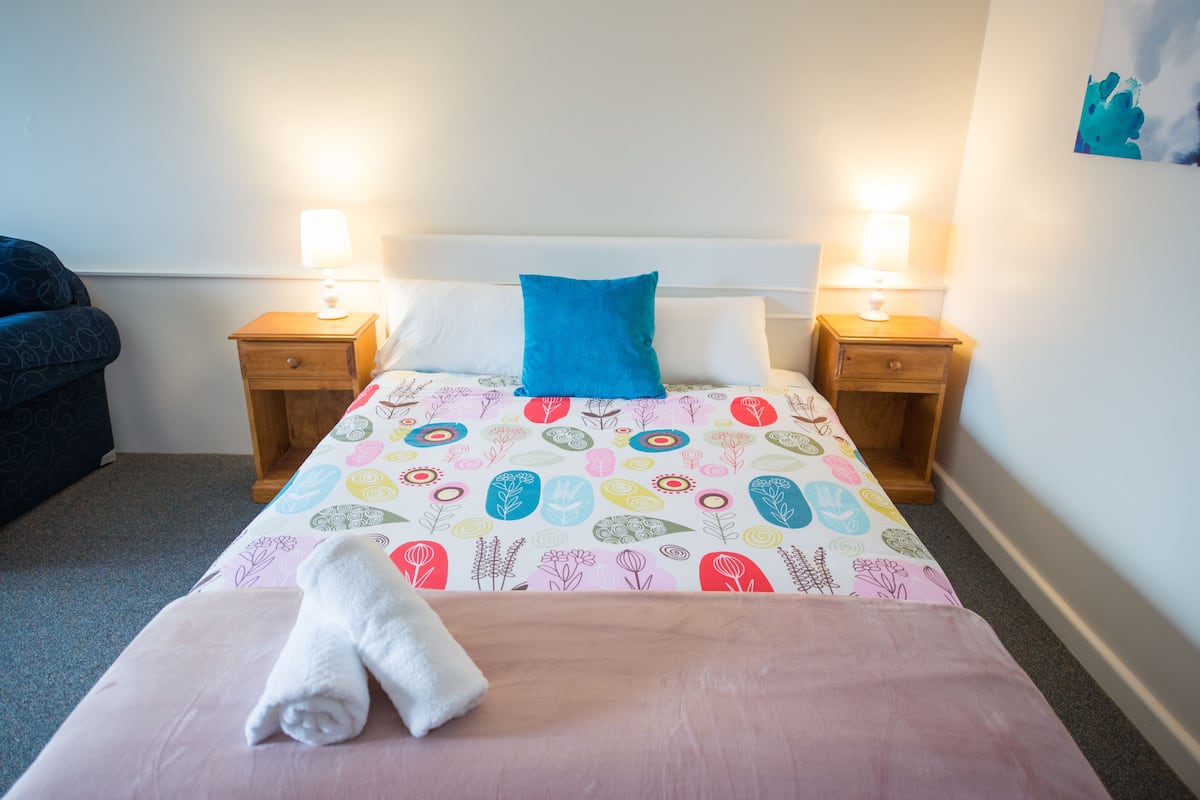 Una cama bien hecha con una colcha floral de colores, dos mesitas de noche con lámparas y un almohador azul.