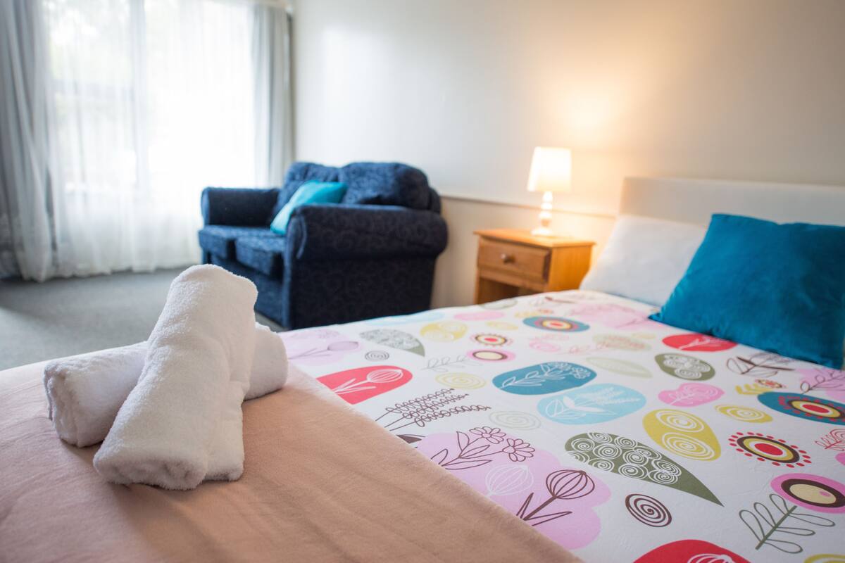 Una habitación de hotel con cama, una silla, una mesita de noche y un ventanal con cortinas.