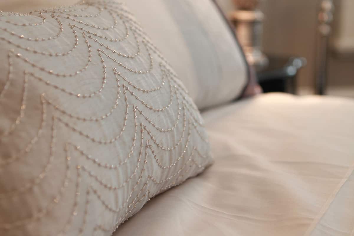A close-up of a bed with a decorative pillow and a lamp in the background.
