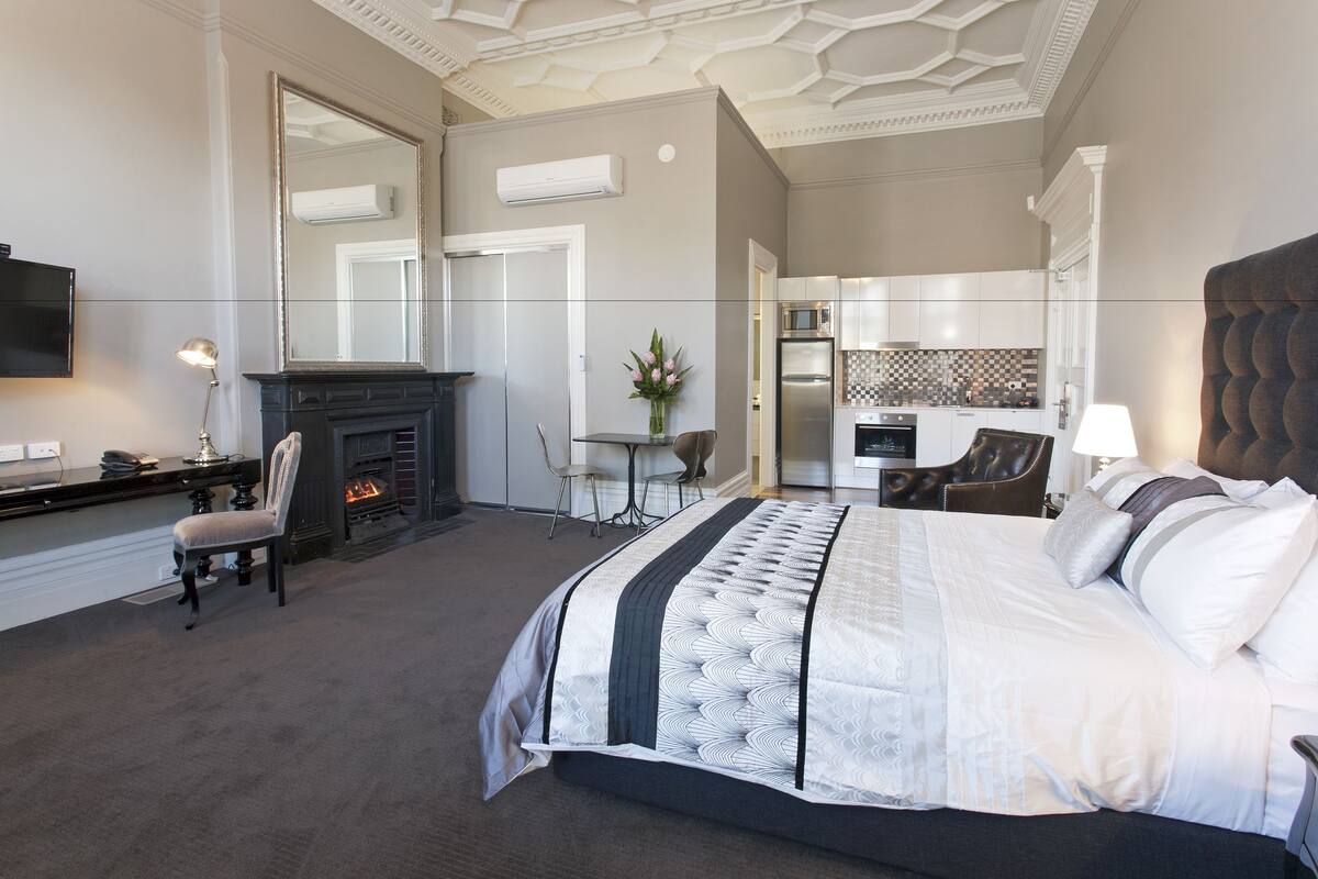 A modern hotel room with a large bed, a fireplace, a dining area, and a kitchenette.