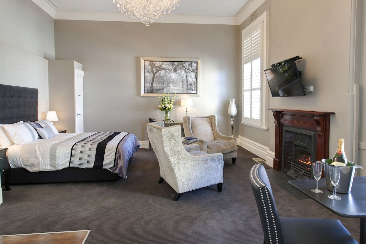 A bedroom with a bed, two armchairs, a table with glasses and a bottle, a fireplace, and a TV mounted on the wall.