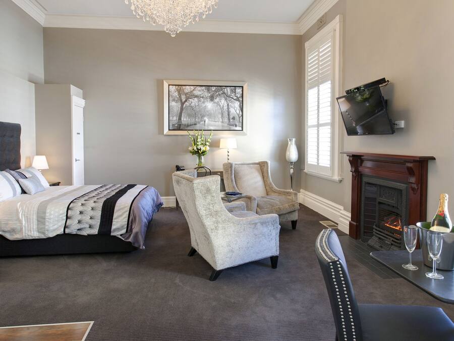A bedroom with a bed, two armchairs, a table with glasses and a bottle, a fireplace, and a TV mounted on the wall.