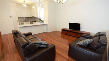A modern living room with two leather sofas, a wooden floor, a flat-screen TV, and a kitchen area with white cabinets and a stone backsplash.