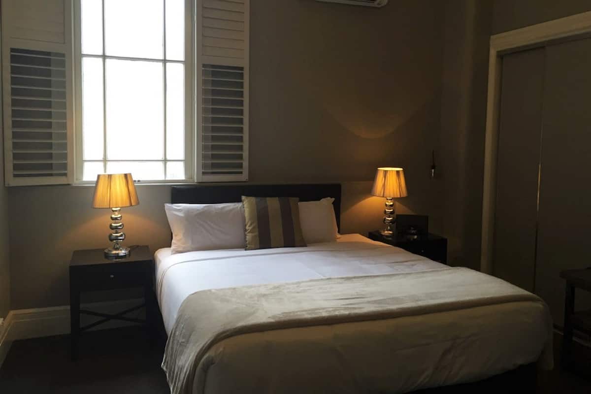 A bedroom with a bed, two bedside lamps, a window with shutters, an air conditioning unit, and a door.