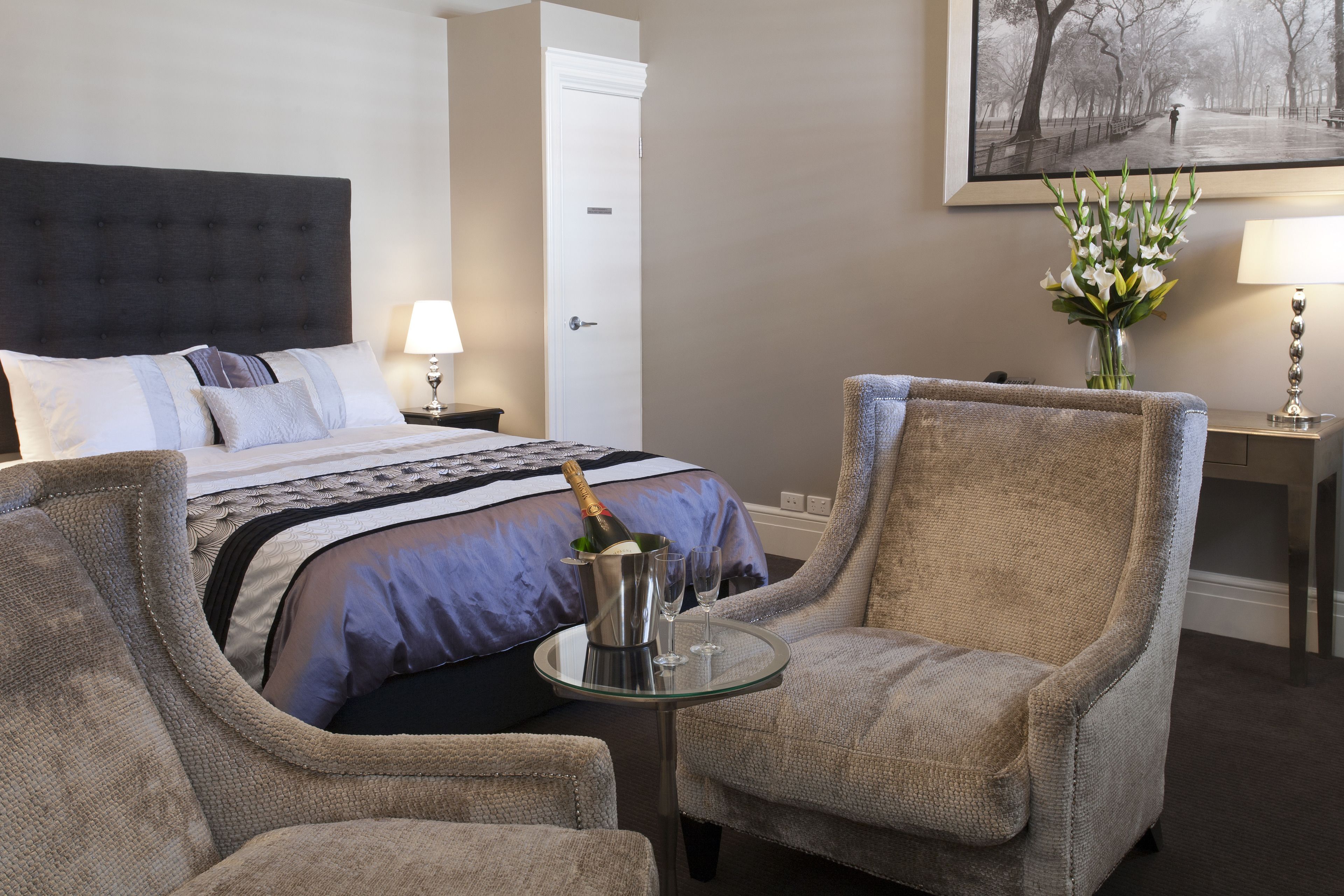 Habitación de hotel con una cama, dos sillones, una mesita con una botella y vasos, un florero con flores y un cuadro en la pared.