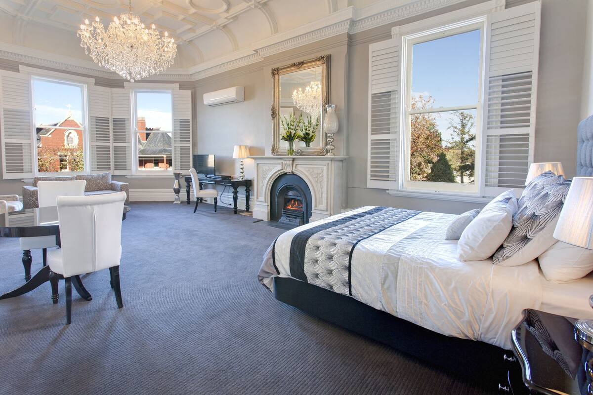 A bedroom with a large bed, a desk, a chair, a fireplace, and large windows.