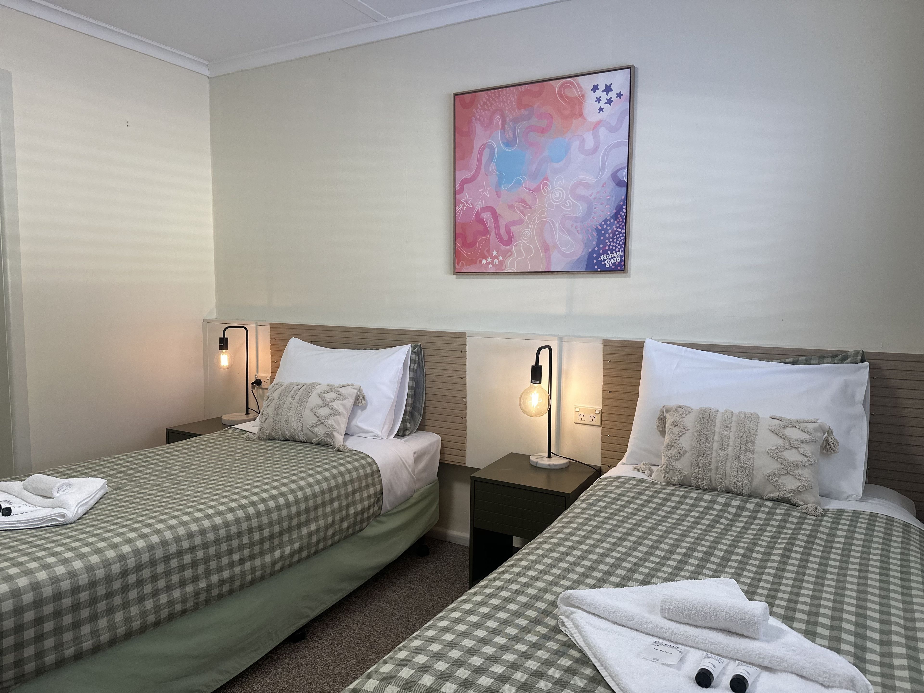 A hotel room with two beds, each with a checkered bedspread and pillows, a bedside table with a lamp, and a wall-mounted abstract painting.