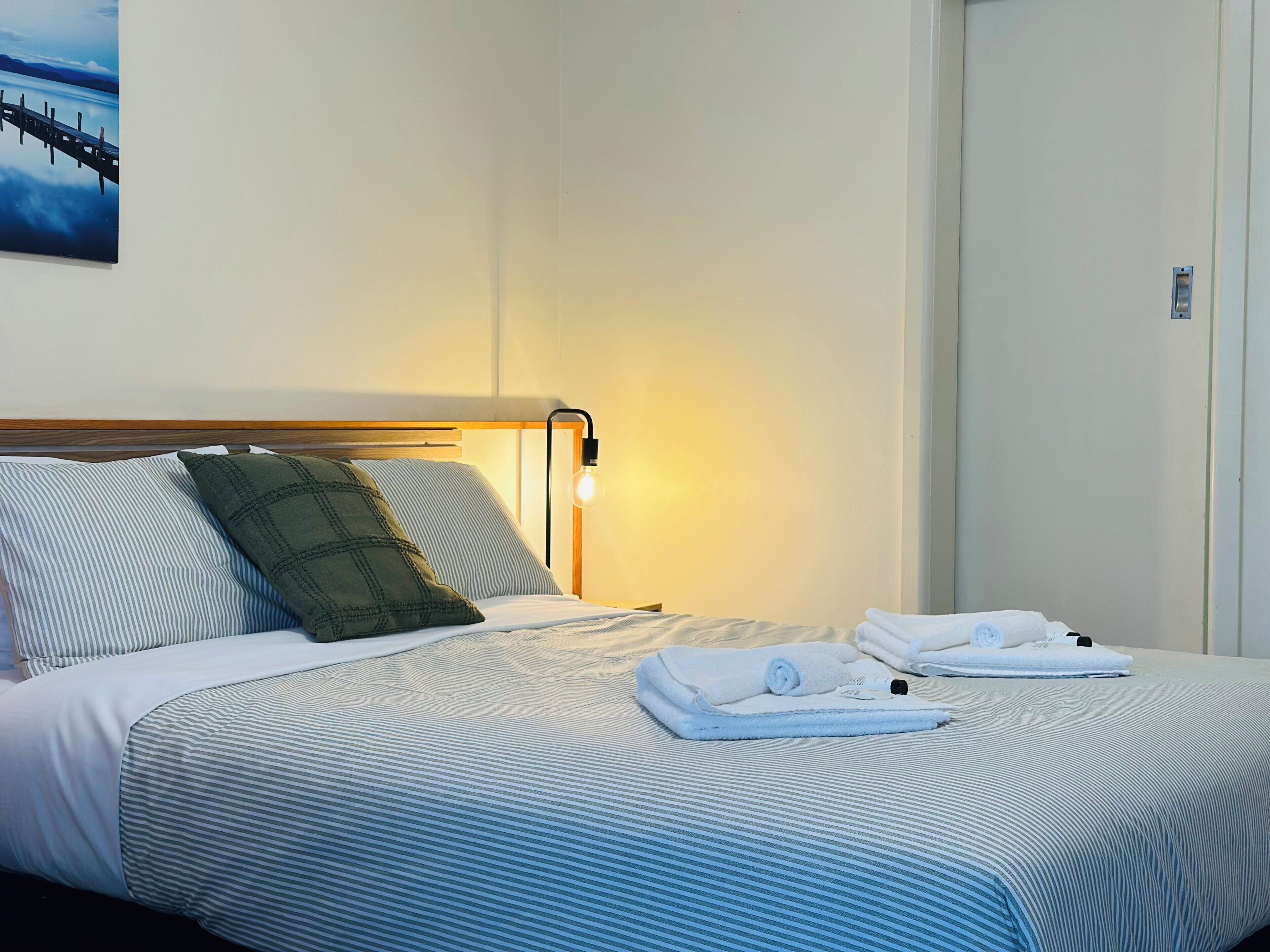 A hotel room with a bed, pillows, a nightstand, a lamp, and a painting on the wall.