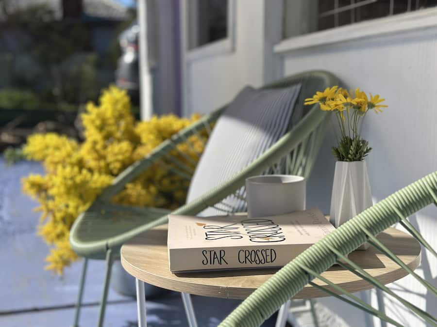 Una silla con cojín, un libro titulado 'STAR CROSSED', un florero con flores amarillas y una manguera verde sobre una mesa afuera de un edificio.