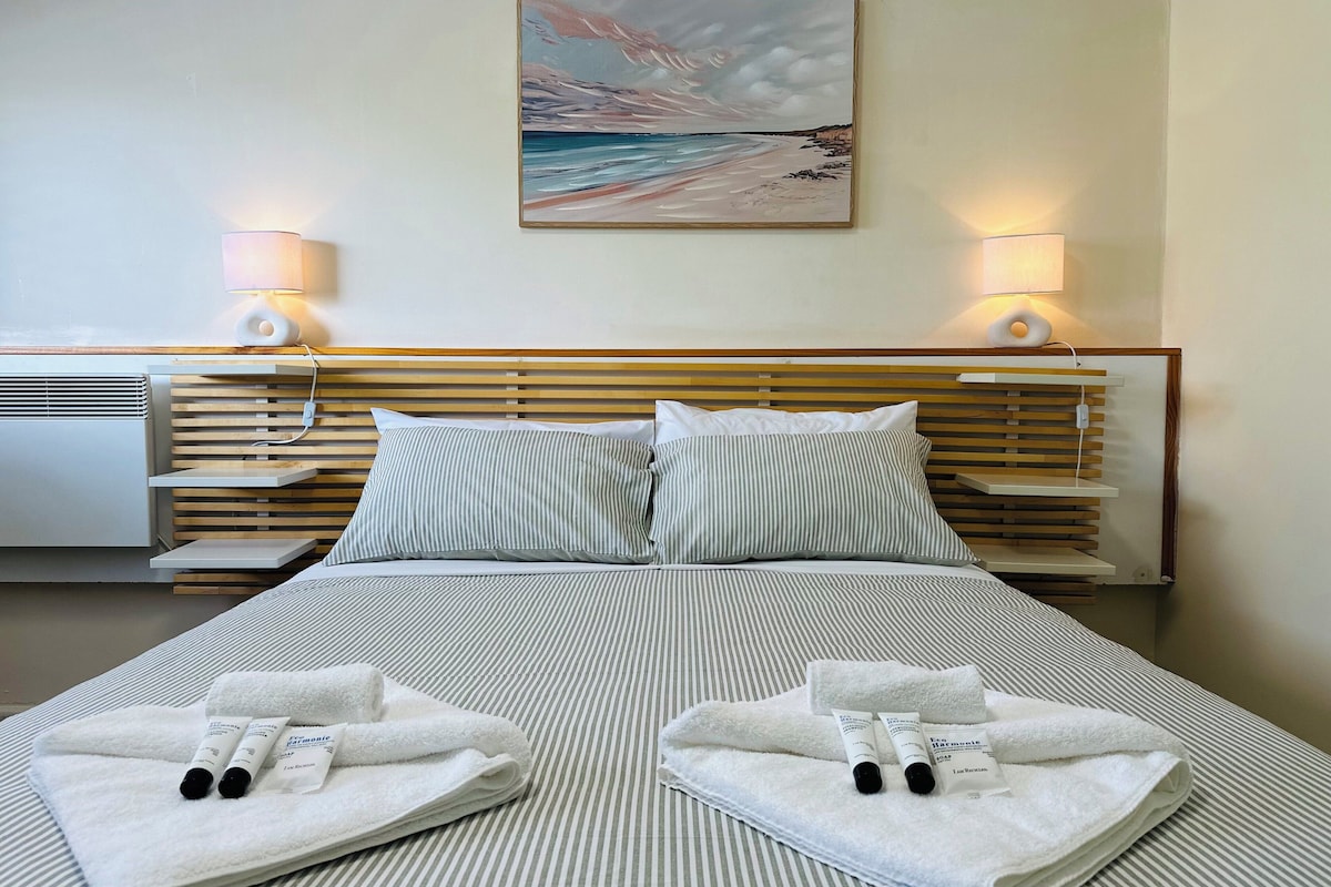 Habitación de hotel con cama, dos lámparas de noche, un cuadro en la pared y una cama bien hecha con toallas y artículos de aseo.