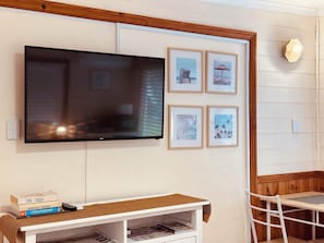 A flat-screen TV mounted on a wall, a small white table with a woven table runner, and a white chair.