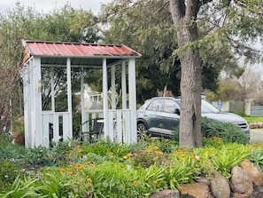 A gazebo with a metal roof and white walls, surrounded by a garden with various plants and flowers.