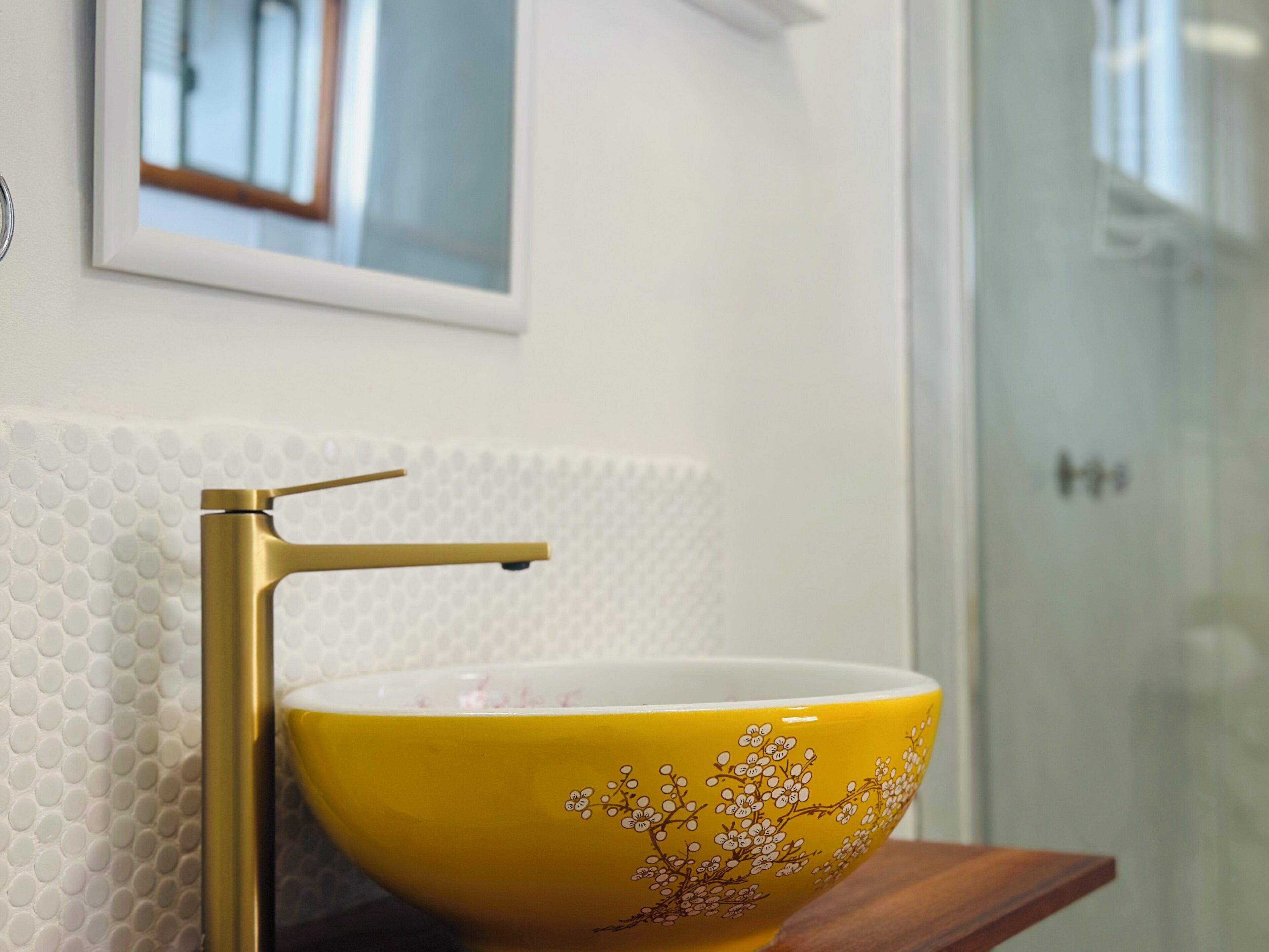 Een badkamer met een geel wasbakje, een gouden kraan en een witte, gestructureerde muur.