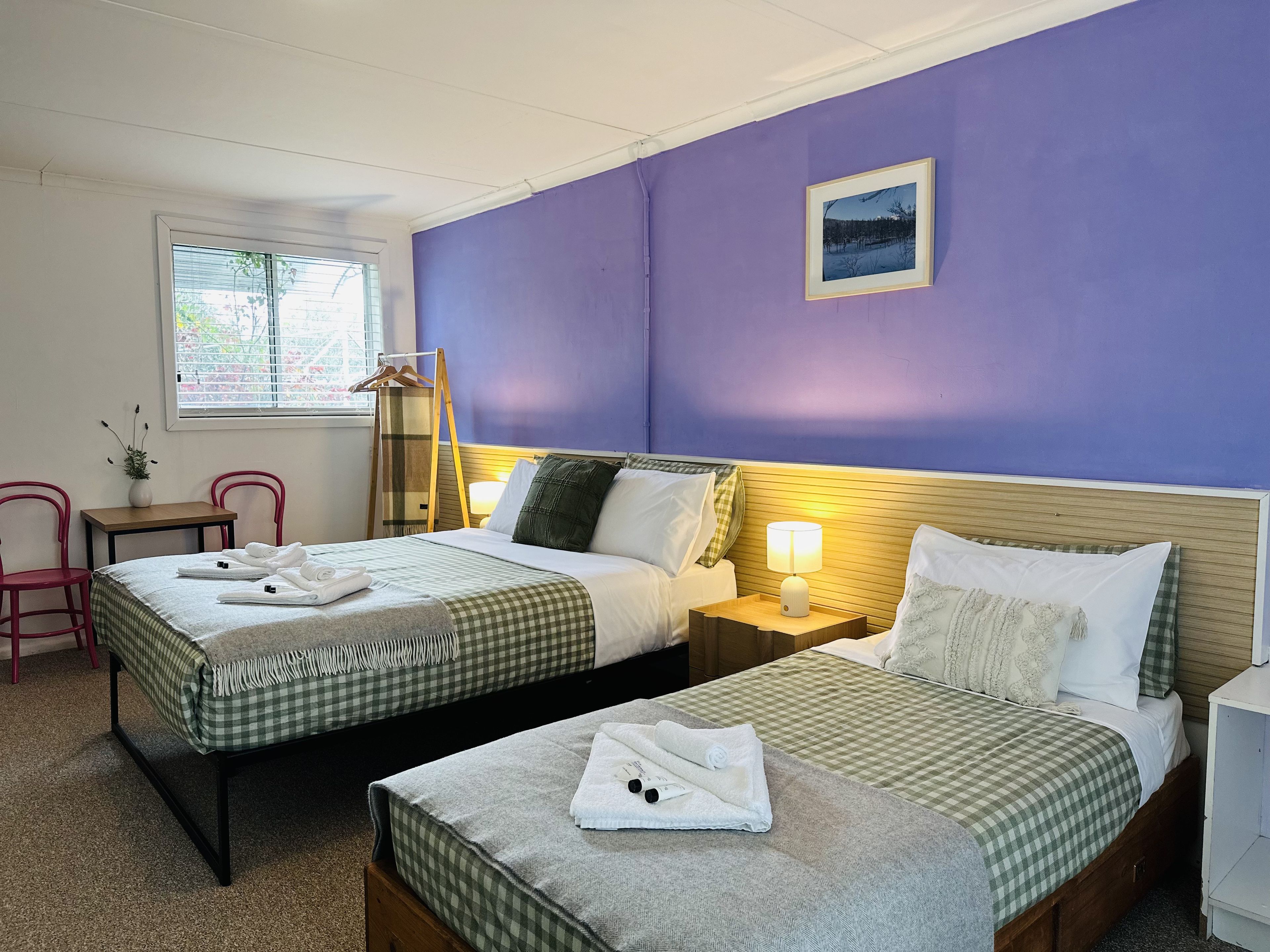 A hotel room with two beds, a window with blinds, a bedside table with a lamp, and a wall-mounted picture.