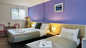 A hotel room with two beds, a window with blinds, a bedside table with a lamp, and a wall-mounted picture.