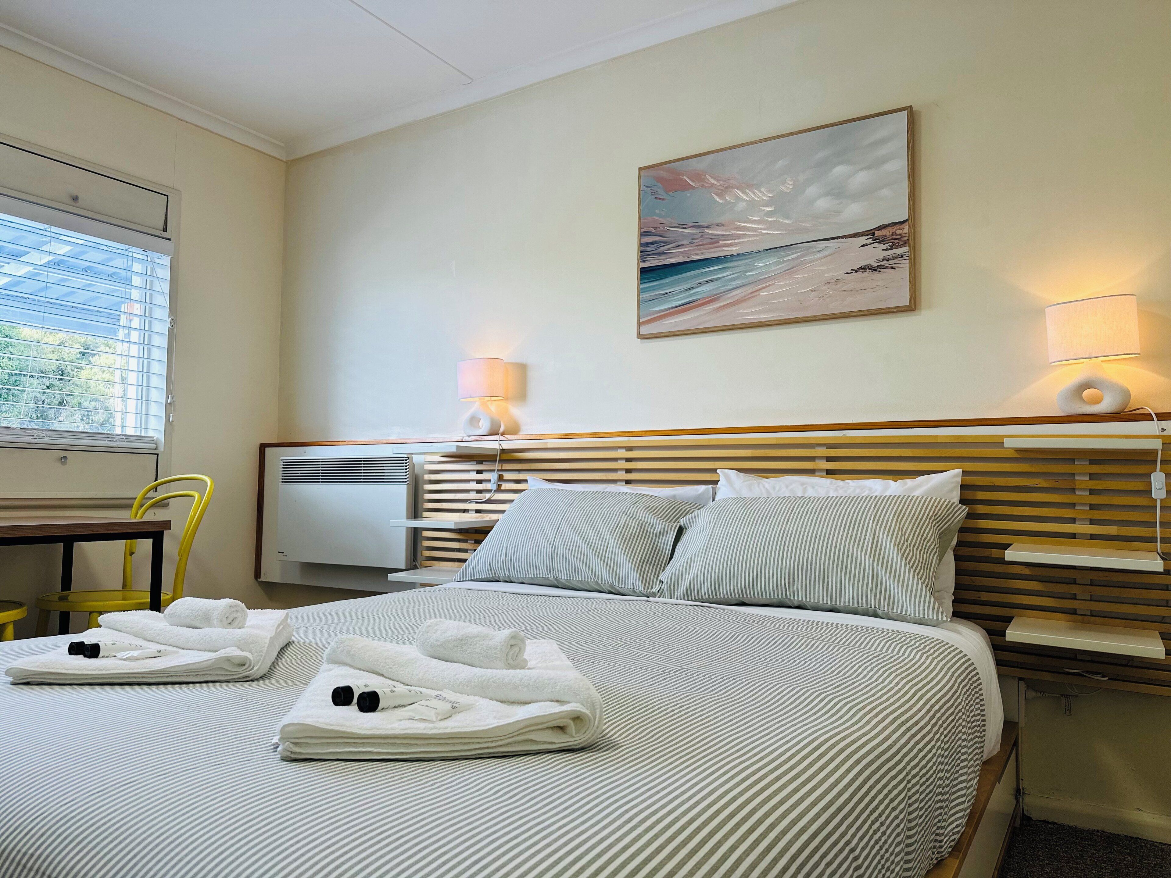 A hotel room with a bed, a painting, a desk, and a chair.