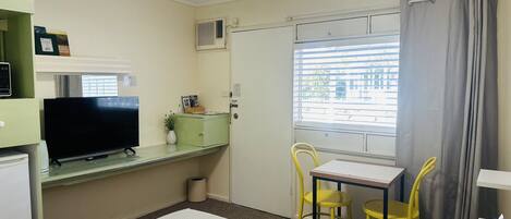 A hotel room with a bed, a desk with a television, a chair, a small table, and a window with curtains.