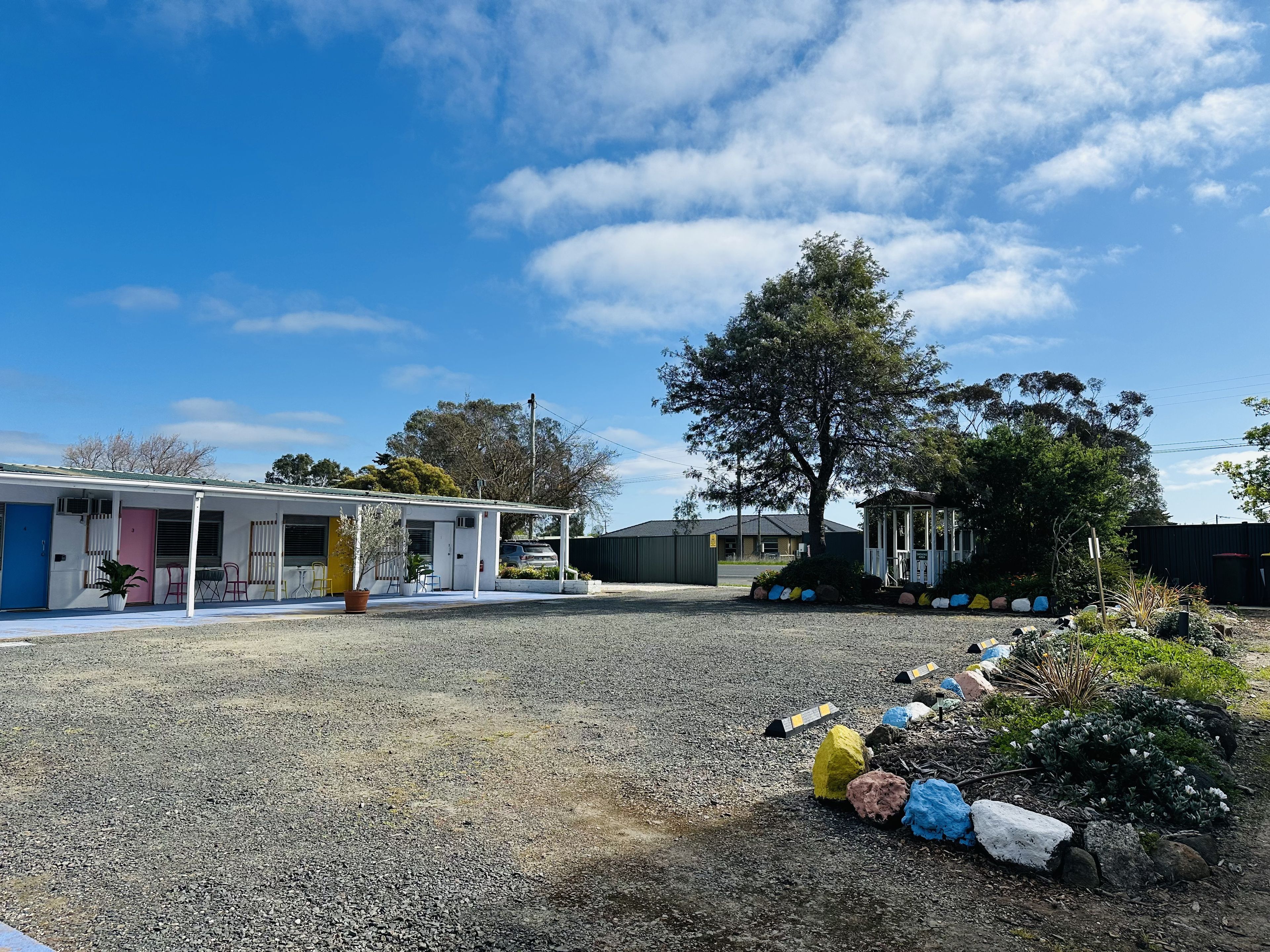 A motel with a covered outdoor seating area, a large tree, and a gravel parking lot.