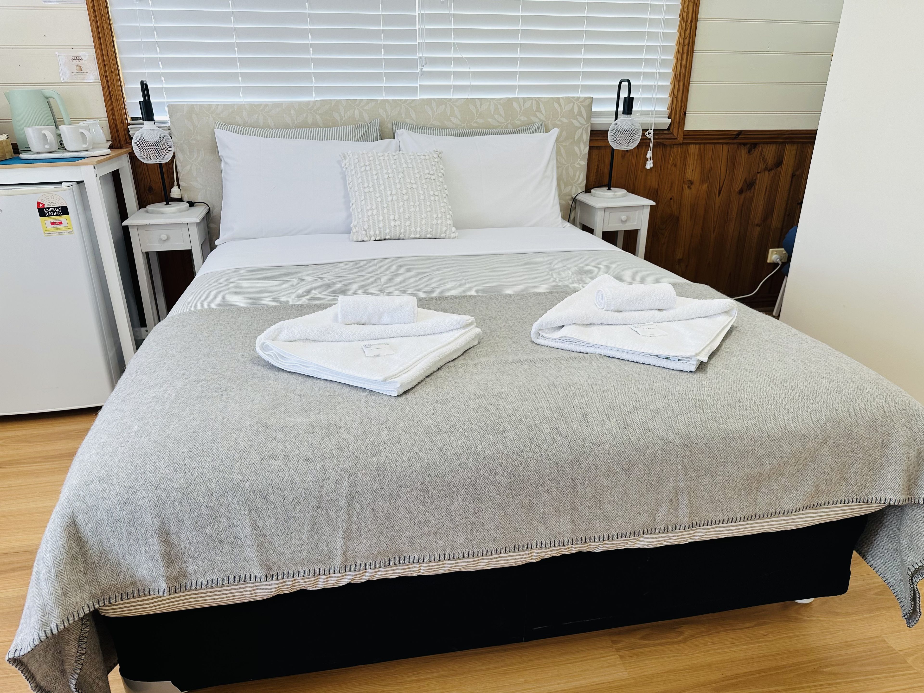 A neatly made bed with a grey bedspread, two folded white towels, and a bedside table with a lamp.