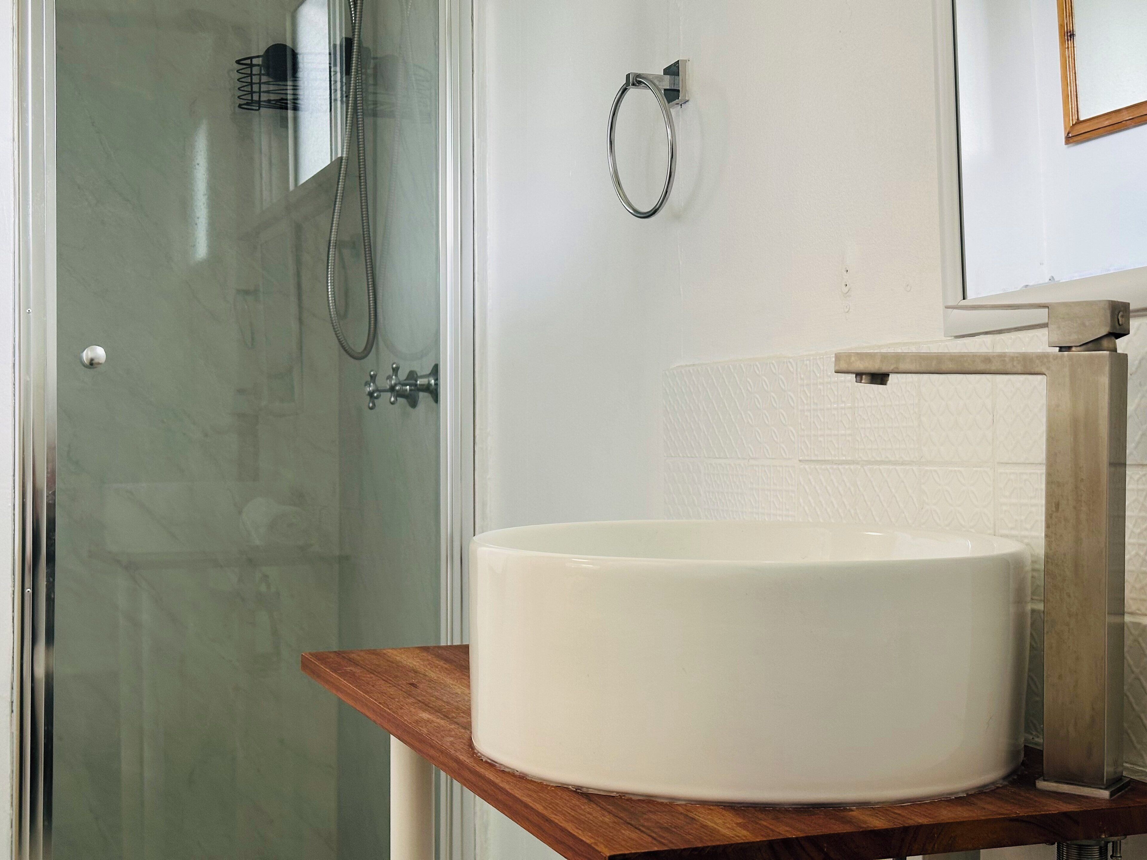 A bathroom with a glass shower enclosure, a white sink on a wooden shelf, and a towel ring on the wall.