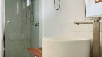 Un baño con cabina de ducha de vidrio, un lavamanos blanco sobre una repisa de madera y un aro para toallas en la pared.
