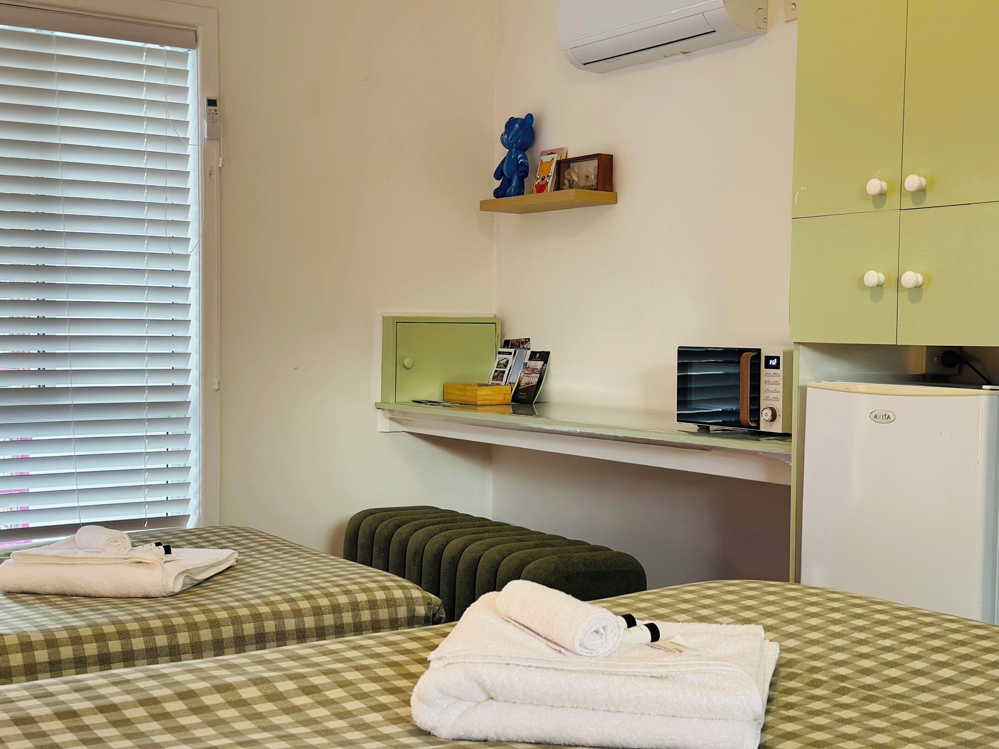 A room with two beds, a small kitchenette, and a window with blinds.