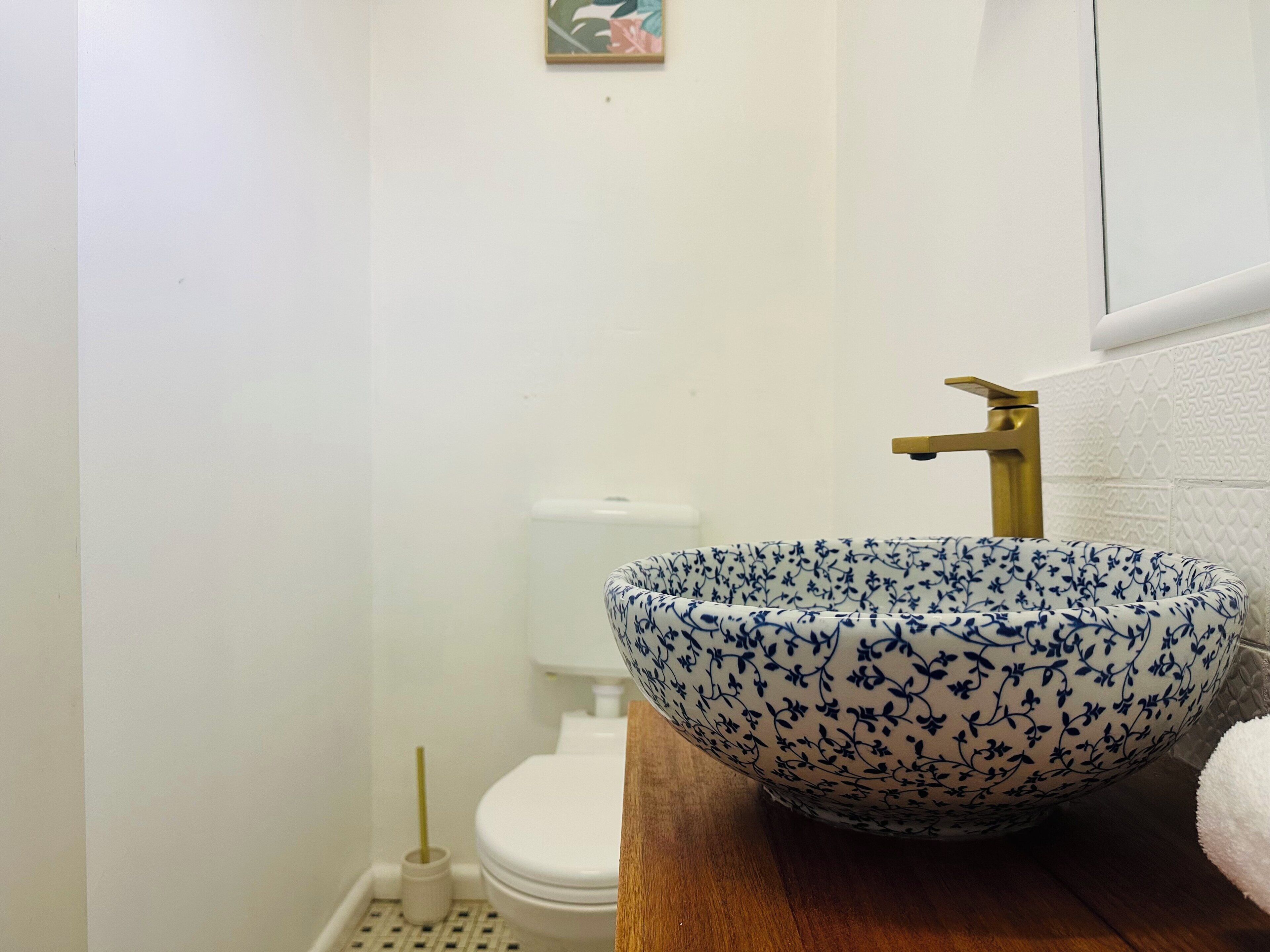 Une salle de bain avec des toilettes blanches, un lavabo en bois orné d’un motif floral bleu et un robinet doré.