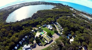 An aerial view of a coastal area with a large body of water, dense greenery, and a cluster of buildings with visible parking areas. - Robe Holiday Park (Robe)