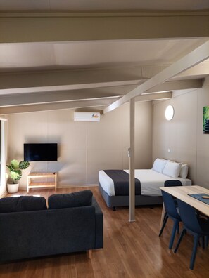 A modern, well-lit room with a wooden floor, a sofa, a bed, a desk with chairs, a TV, and a potted plant. - Robe Holiday Park (Robe)