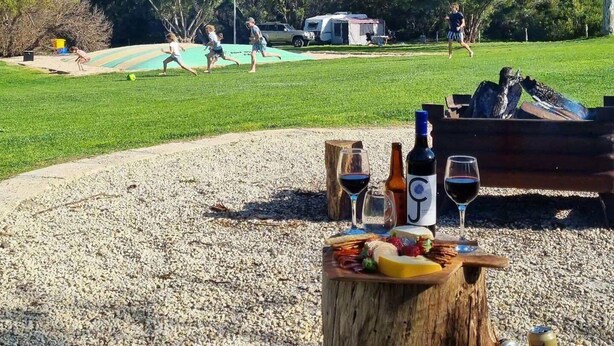A picnic setup with food and drinks on a wooden stump, surrounded by a gravel area with a grassy field and trees in the background.