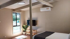 A modern hotel room with a wooden floor, a bed with a dark headboard, a desk with a chair, a small table with a plant, and a large window with a view of greenery.