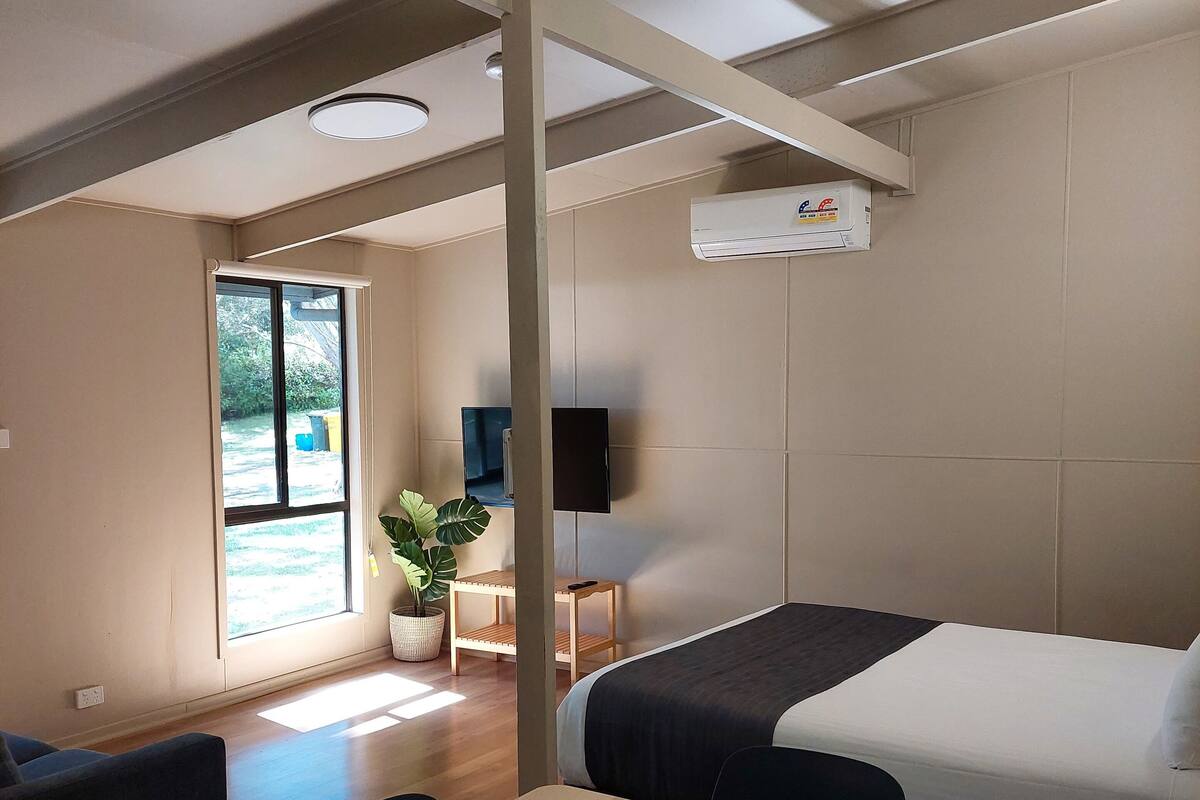 A modern hotel room with a wooden floor, a bed with a dark headboard, a desk with a chair, a small table with a plant, and a large window with a view of greenery.