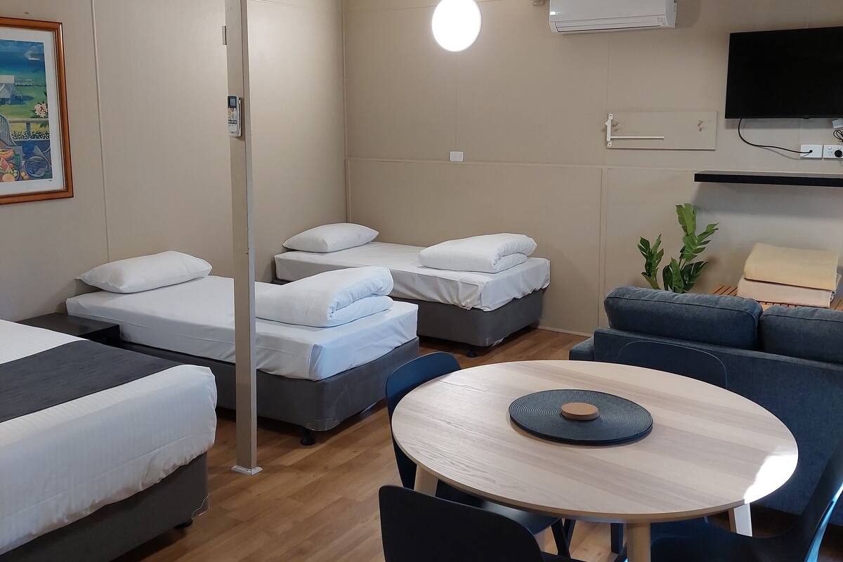 A modern, well-lit room with a round wooden table, blue chairs, and a sofa. There are four beds with white bedding, a flat-screen TV mounted on the wall, and an air conditioning unit.