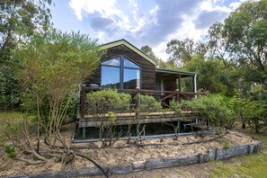 A cabin with a large window, wooden deck, and surrounded by trees. - Wisteria Cottage and Cabins (Wyberba)