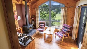 Ein gemütliches Blockhaus-Interieur mit Holzofen, Karokreuz-Stühlen, einem Tisch und einem großen Fenster mit Blick auf eine grüne Landschaft.
