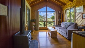 Ein gemütliches Blockhaus-Interieur mit Holzbalkendecke, Kamin, einer Couch und einem großen Fenster mit Blick ins Freie.