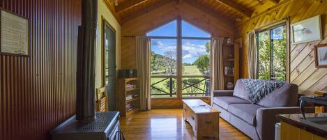 Interior acogedor de una cabaña con techo de madera, chimenea, sofá y un amplio ventanal con vistas al exterior.
