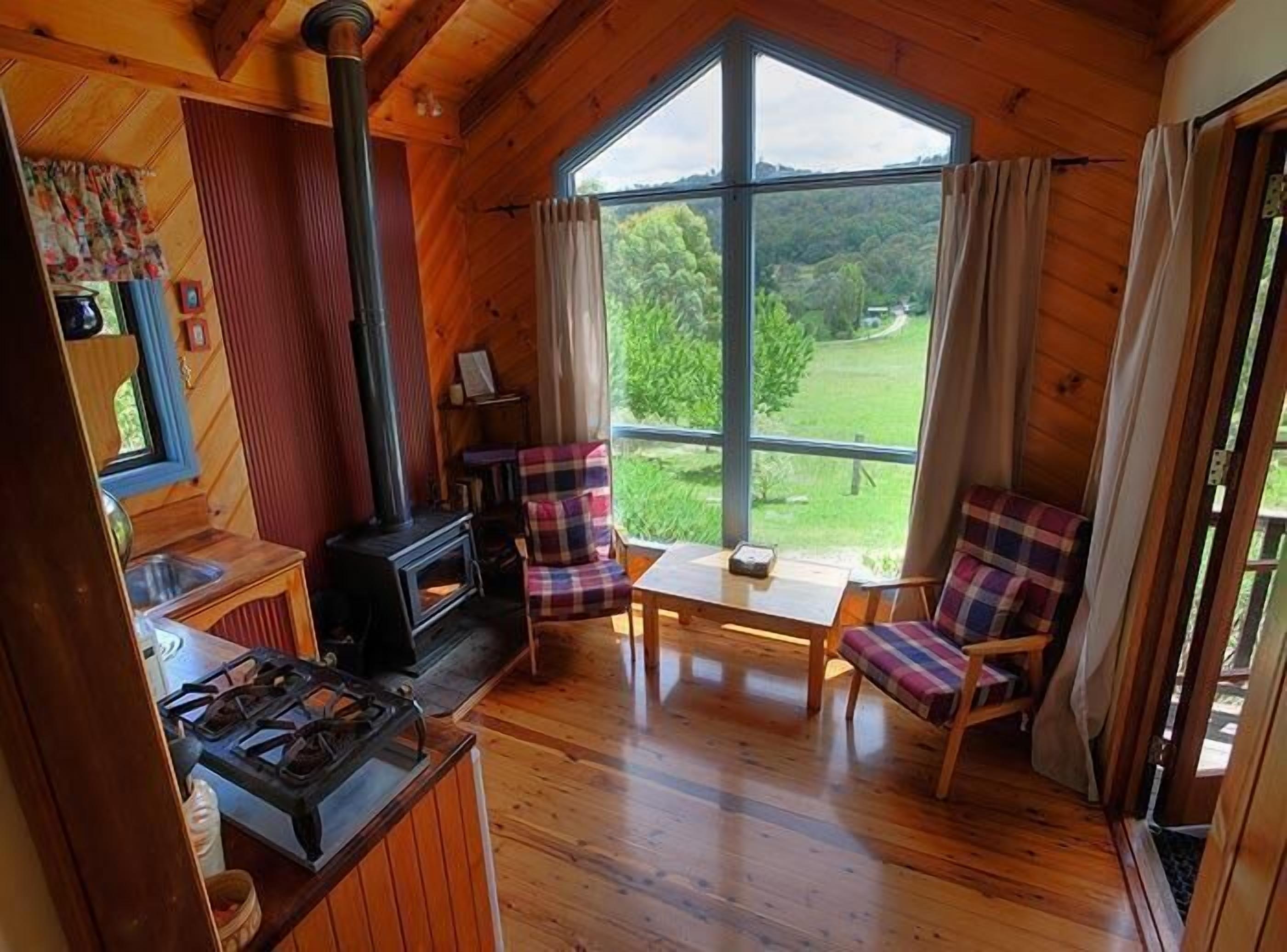A cozy cabin with a wood stove, plaid chairs, and a large window offering a view of the forest.