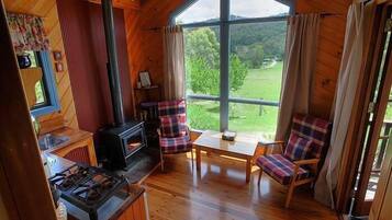 A cozy cabin with a wood stove, plaid chairs, and a large window offering a view of the forest.